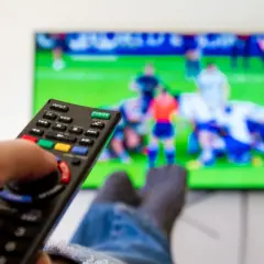 Dłoń trzymająca pilot do telewizora, na ekranie mecz piłki nożnej.