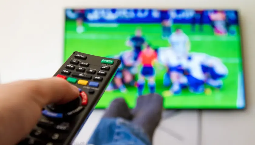 Dłoń trzymająca pilot do telewizora, na ekranie mecz piłki nożnej.