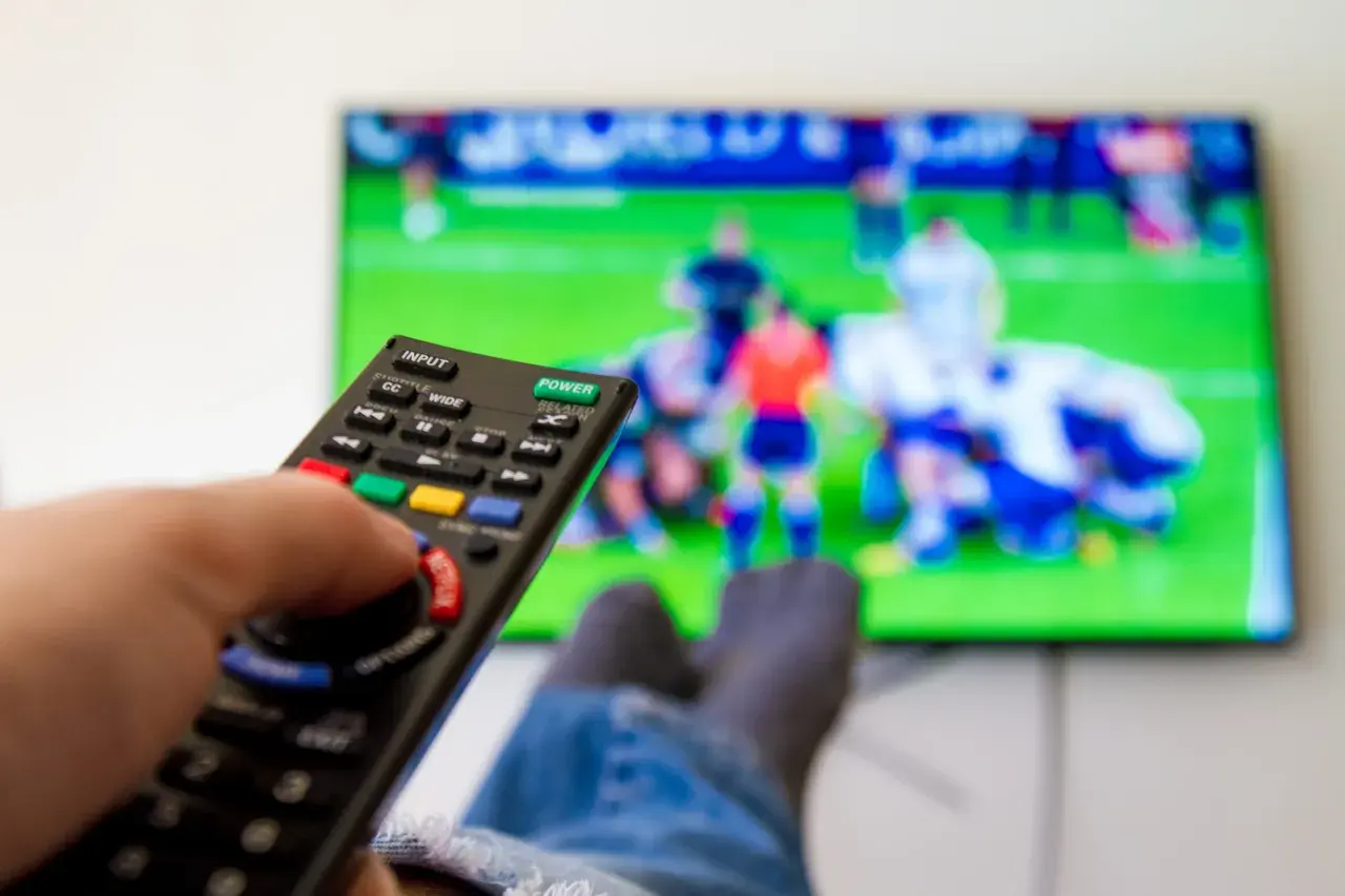 Dłoń trzymająca pilot do telewizora, na ekranie mecz piłki nożnej.